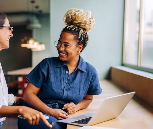 Two young women smiling and talking while working together at a laptop Two women are seated in a bright indoor setting, engaging in conversation near a laptop. They appear to be sharing ideas and enjoying their discussion while wearing casual attire.